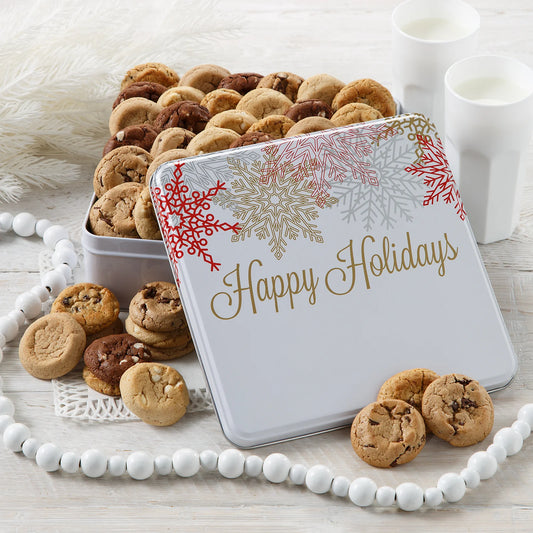 A holiday themed cookie tin that says "Happy Holidays" with colorful snowflakes filled with Mrs. Fields Nibblers bite-sized cookies on a white background with a glass of milk.