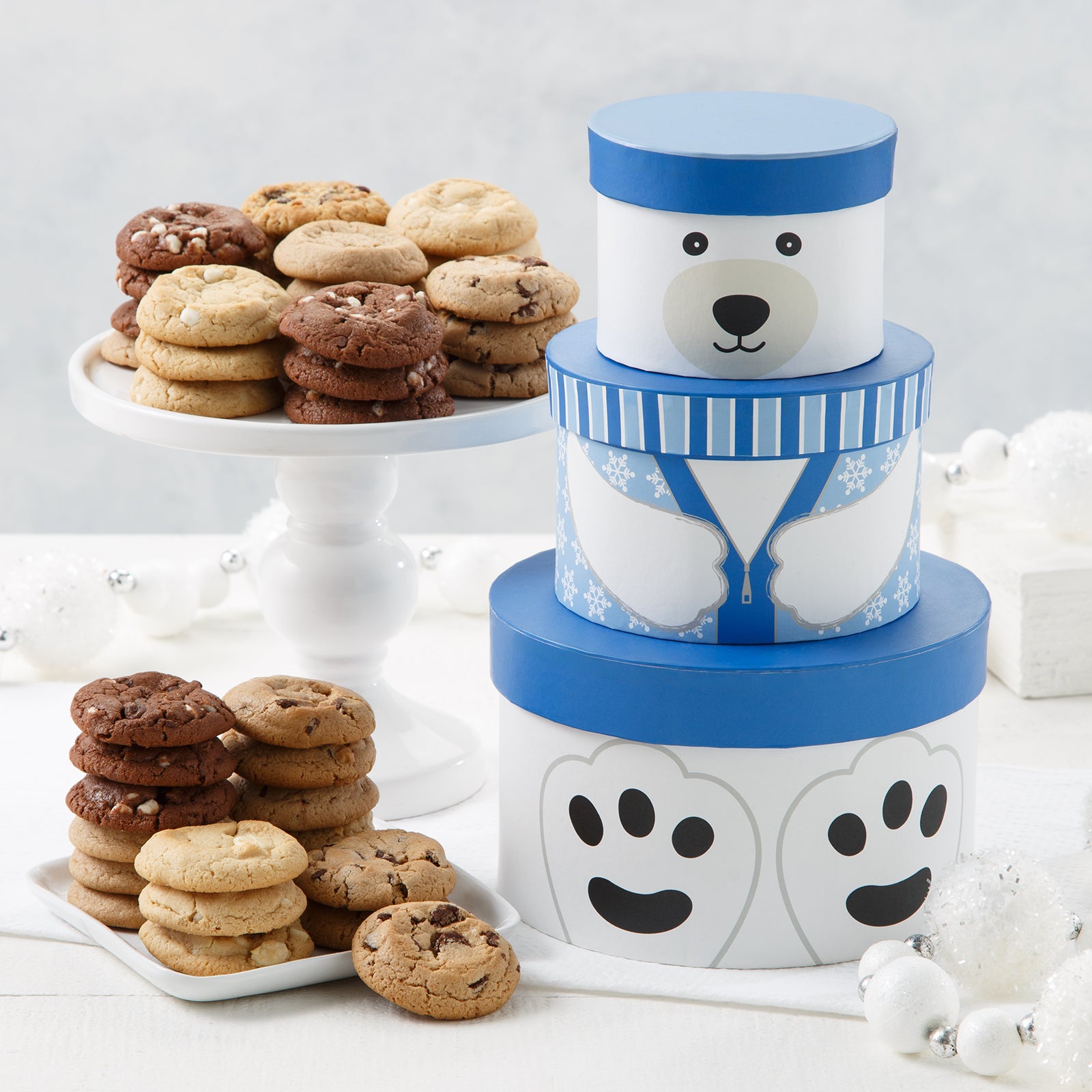 A three-tiered blue and white tower of round boxes that make a polar bear when stacked together surrounded by an assortment of Mrs. Fields cookies in five flavors including chocolate chip and cinnamon sugar on a white surface with white Christmas ornaments.