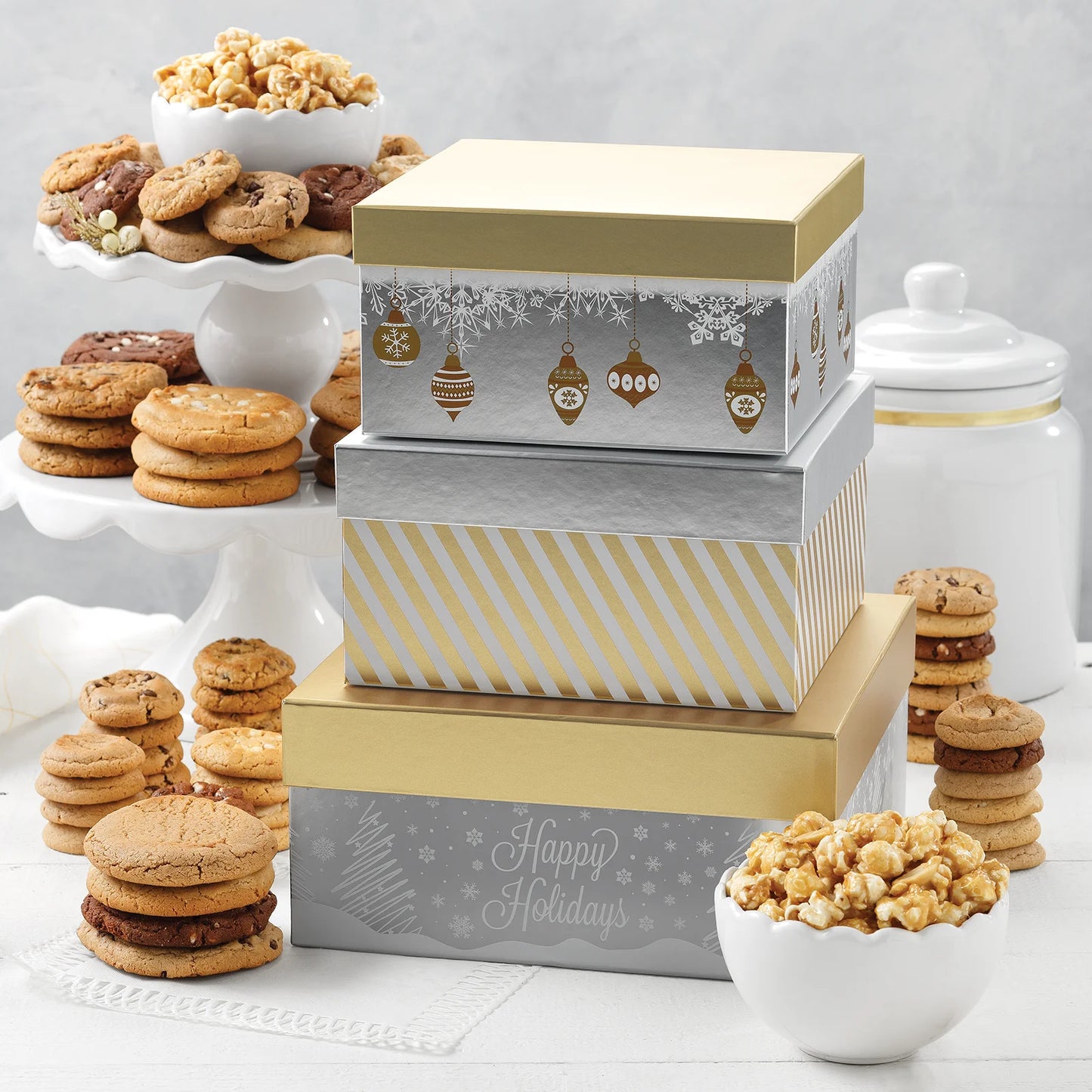 A three-tiered holiday themed tower of silver and gold boxes surrounded by an assortment of Mrs. Fields cookies in five flavors including chocolate chip and cinnamon sugar, and toffee nut popcorn on a white surface.