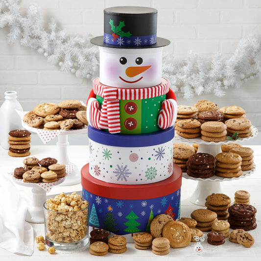 A five-tiered tower of boxes that makes a snowman figure wearing a top hat and knitted scarf and mittens, surrounded by a large assortment of Mrs. Fields cookies and a generous helping of toffee nut popcorn on a white surface with a white garland.