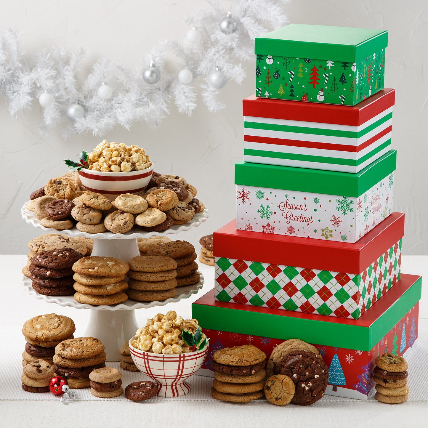 A five-tiered red, green, and white tower of holiday-themed square boxes surrounded by an assortment of Mrs. Fields cookies in five flavors including chocolate chip and white chocolate, and toffee nut popcorn on a white surface with a white garland and white ornaments.
