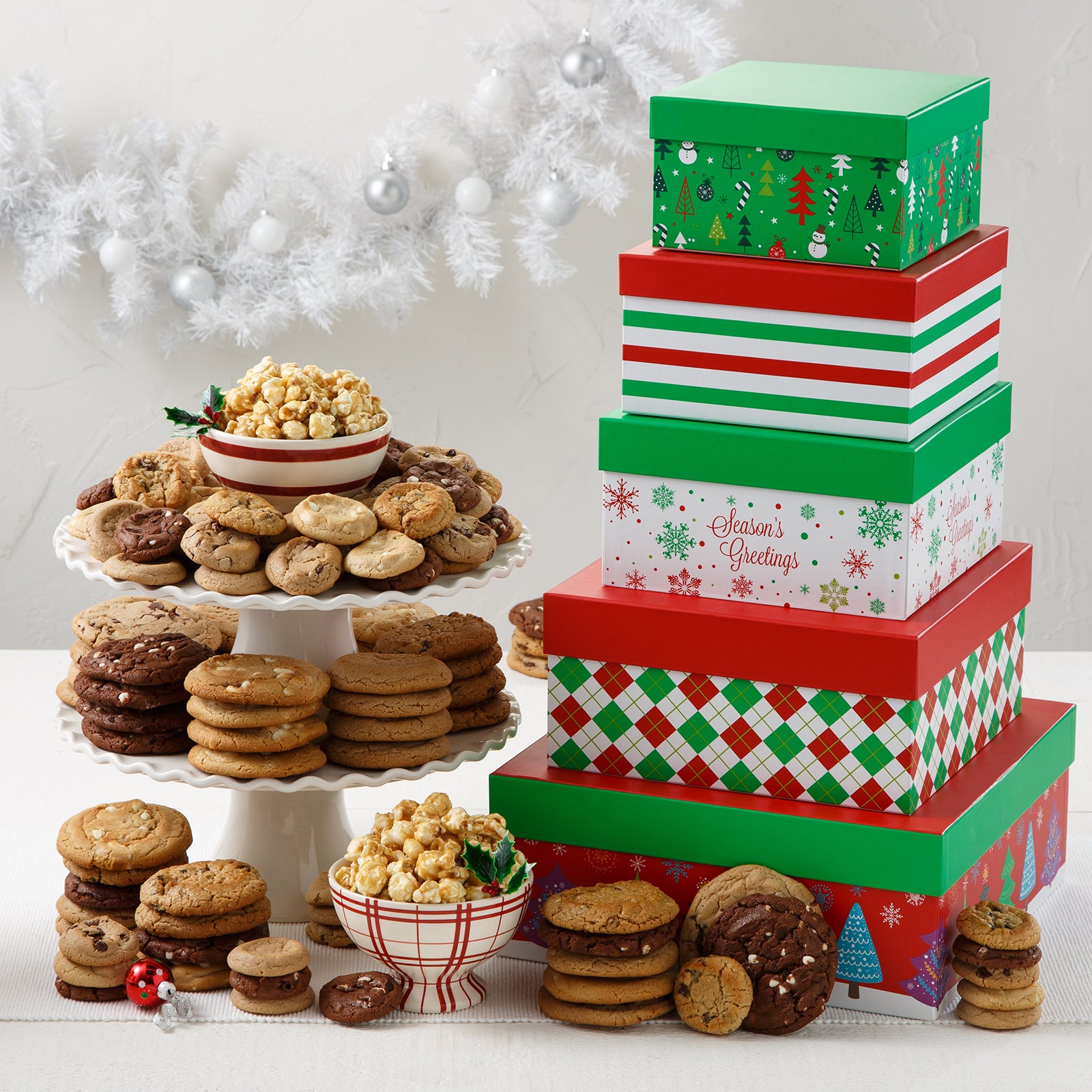 A five-tiered red, green, and white tower of holiday-themed square boxes surrounded by an assortment of Mrs. Fields cookies in five flavors including chocolate chip and white chocolate, and toffee nut popcorn on a white surface with a white garland and white ornaments.