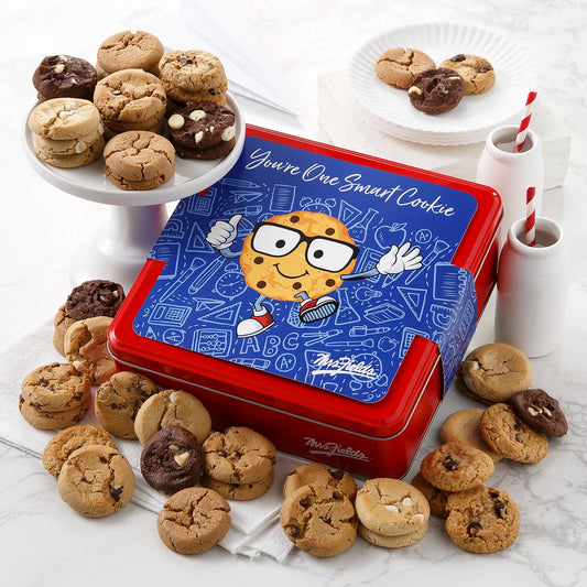 Mrs. Fields signature red cookie tin with a decorative sleeve on it that says "You're One Smart Cookie" with an animated cookie giving a thumbs up, surrounded by an assortment of Nibblers bite-sized cookies on a white surface.