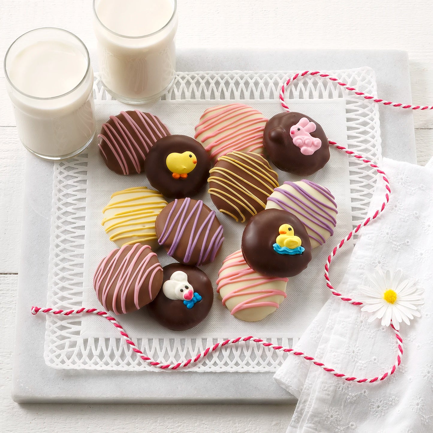 Twelve chocolate covered Nibblers bite-sized cookies with pastel chocolate drizzles and bunny and chick decorations on a white surface with two glasses of milk.