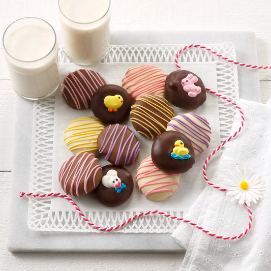 Twelve chocolate covered Nibblers bite-sized cookies with pastel chocolate drizzles and bunny and chick decorations on a white surface with two glasses of milk.