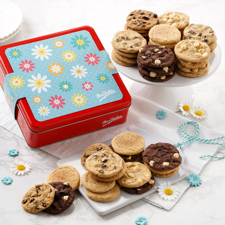 A red Mrs. Fields cookie tin with a pastel floral sleeve on it surrounded by an assortment of Nibblers bite-sized cookies on a white marbled surface with small flowers and blue string.