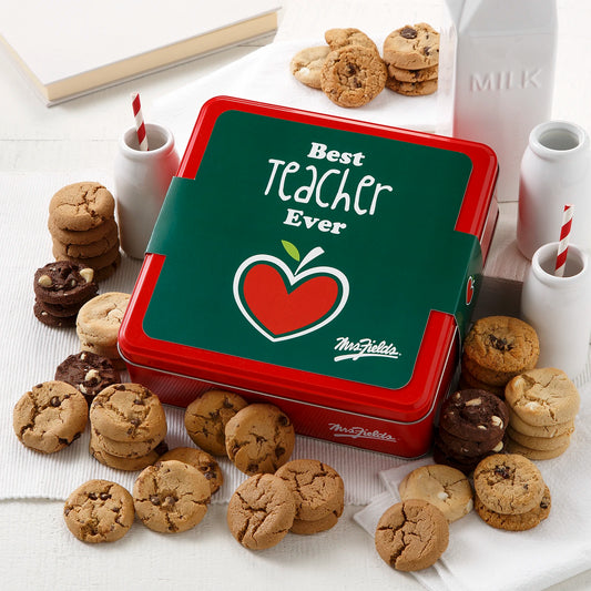 A Mrs. Fields signature red tin with a "Best Teacher Ever" sleeve on it surrounded by an assortment of Nibblers bite-sized cookies on a white surface.