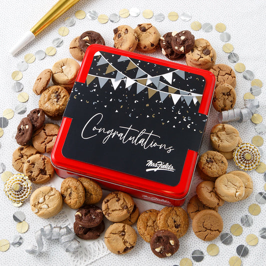 A Mrs. Fields signature red tin with a "Congratulations" sleeve on it surrounded by an assortment of Nibblers bite-sized cookies on a white surface with silver and gold congratulatory decorations.
