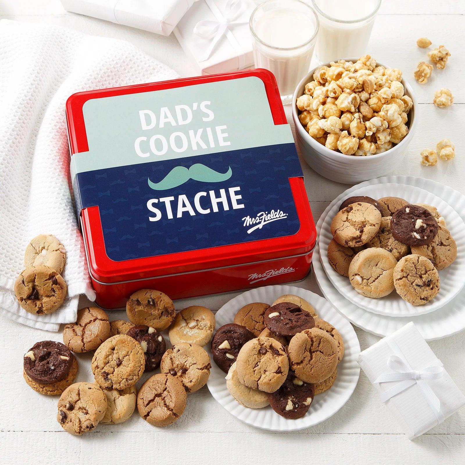Mrs. Fields signature red cookie tin with a Father's Day themed sleeve that says "Dad's Cookie Stache" with a mustache on it surrounded by an assortment of Nibblers bite-sized cookies and toffee nut popcorn on a white surface with two glasses of milk.