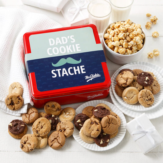 Mrs. Fields signature red cookie tin with a Father's Day themed sleeve that says "Dad's Cookie Stache" with a mustache on it surrounded by an assortment of Nibblers bite-sized cookies and toffee nut popcorn on a white surface with two glasses of milk.