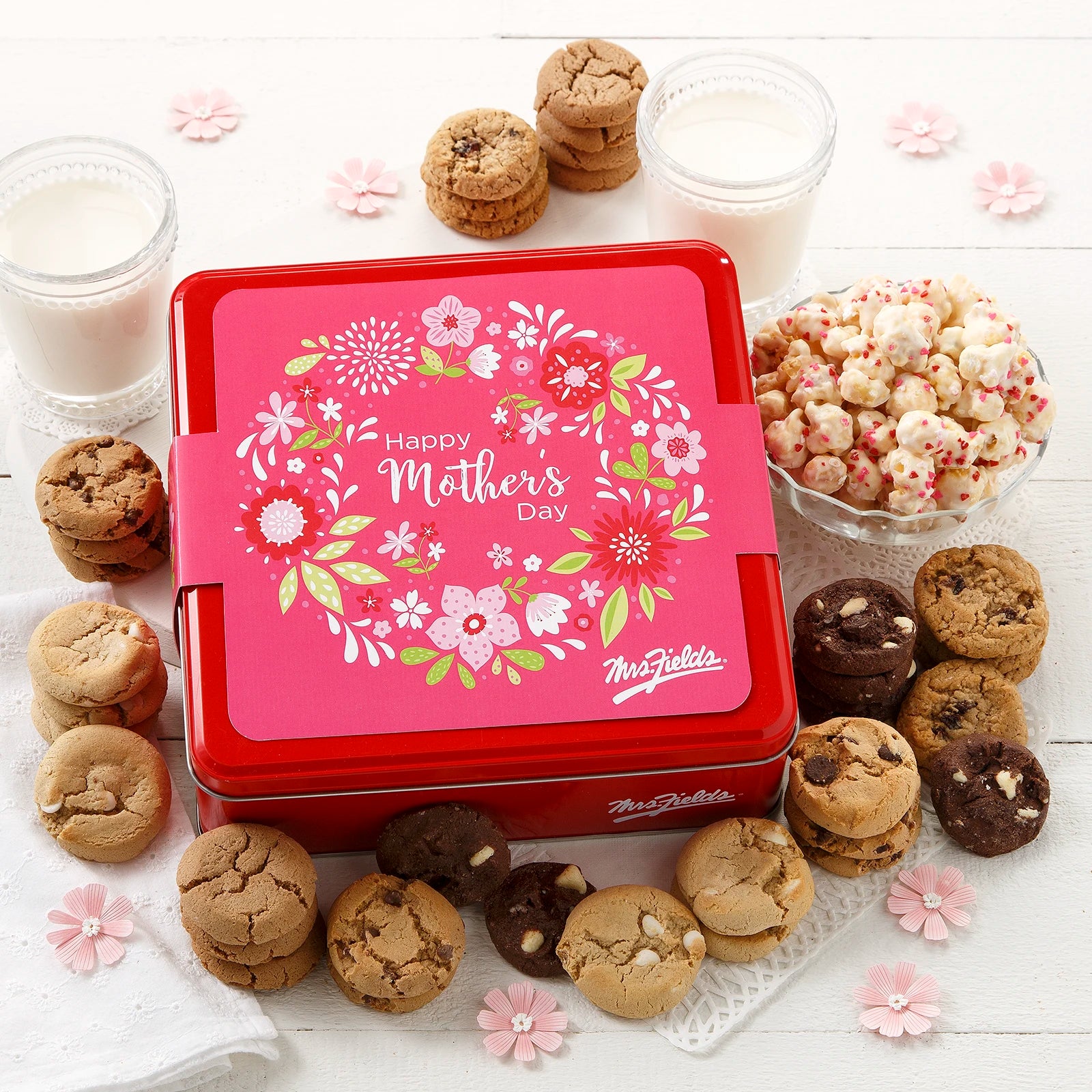 Mrs. Fields signature red cookie tin with a Spring and Mother's Day themed sleeve that says "Happy Mother's Day" with a floral print surrounded by an assortment of Nibblers bite-sized cookies and sweet popcorn with heart sprinkles on a white surface with flower decorations and two glasses of milk.