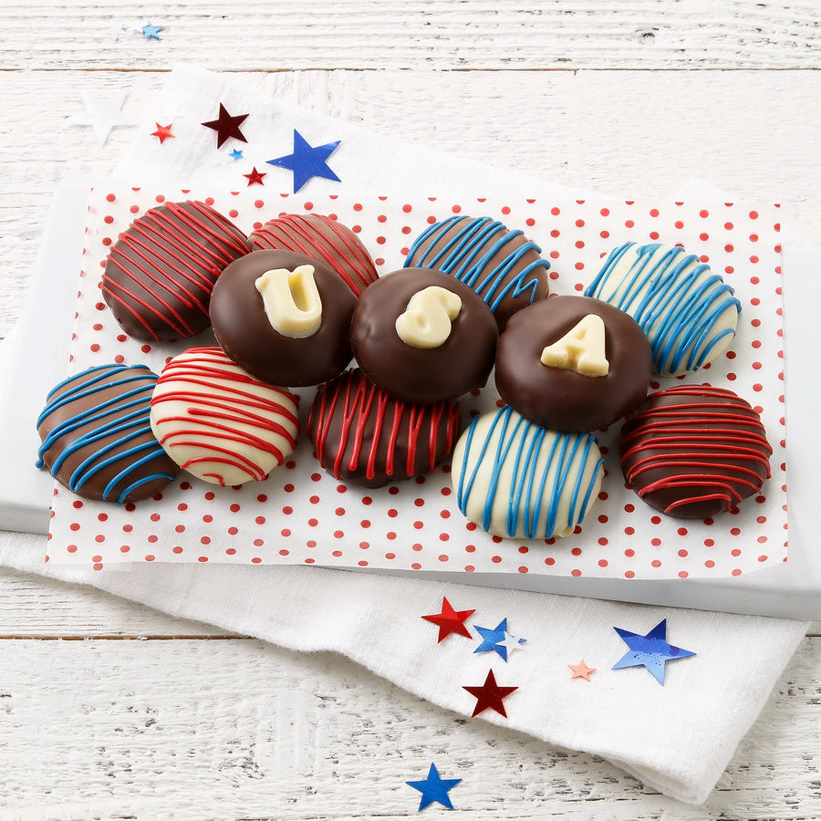 Twelve chocolate covered Nibblers bite-sized cookies decorated in red, white, and blue chocolate drizzles and white chocolate lettering spelling out "USA" on a white surface with star decorations.