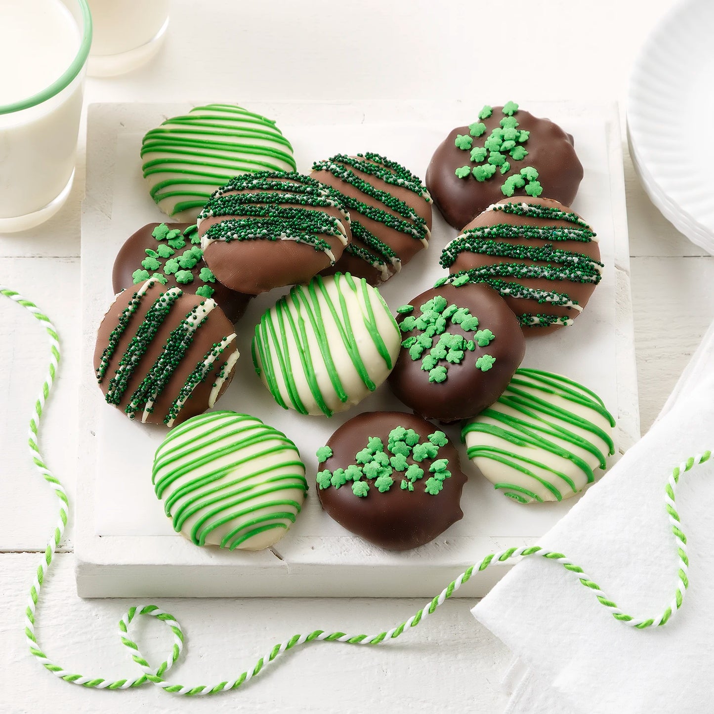 Twelve chocolate covered Nibblers bite-sized cookies with green chocolate drizzles and shamrock sprinkles on a white surface with milk glasses.