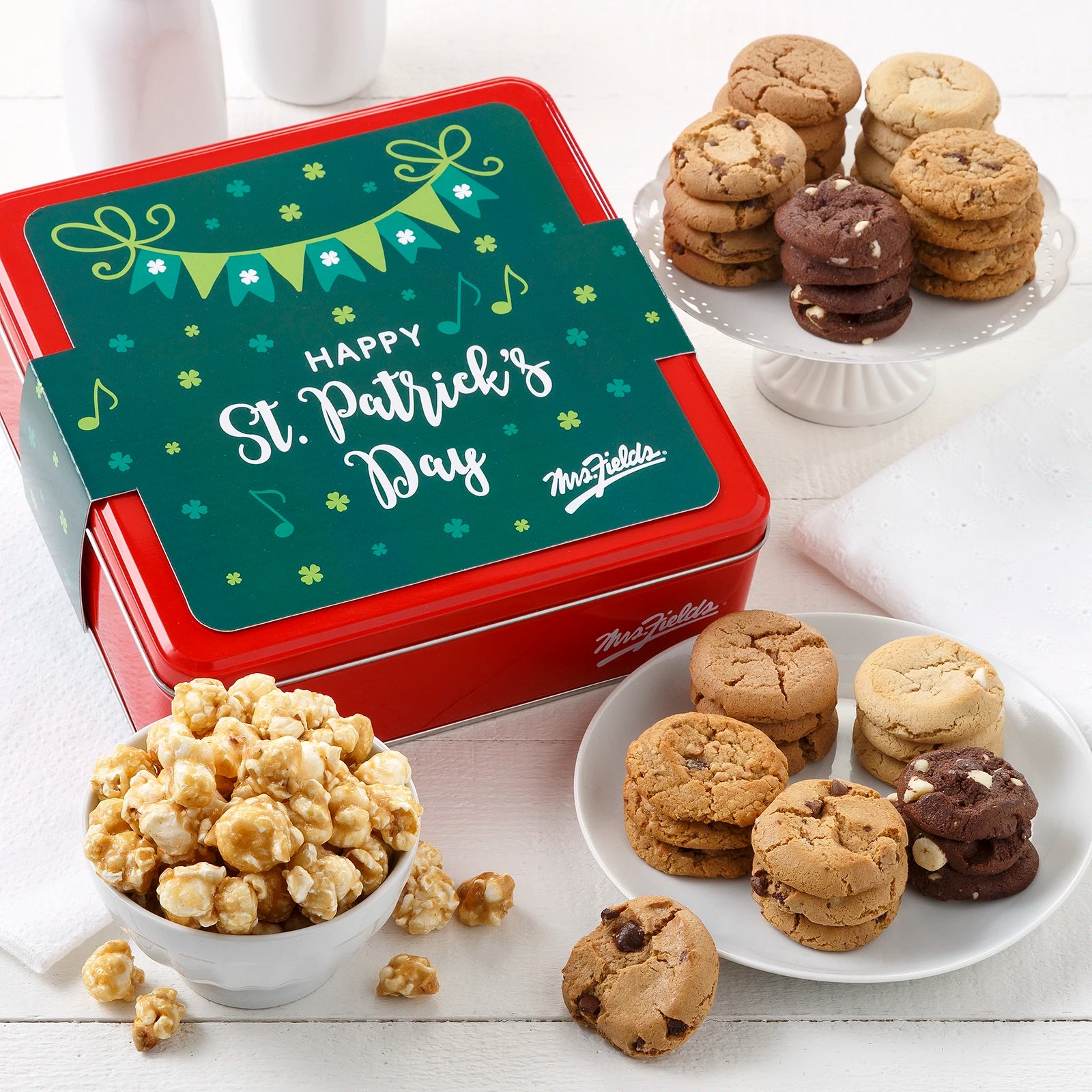 Mrs. Fields red cookie tin with a green clover themed sleeve that says "Happy St. Patrick's Day" surrounded by an assortment of Nibblers bite-sized cookies and toffee nut popcorn on a white surface with white dishes and milk bottles.