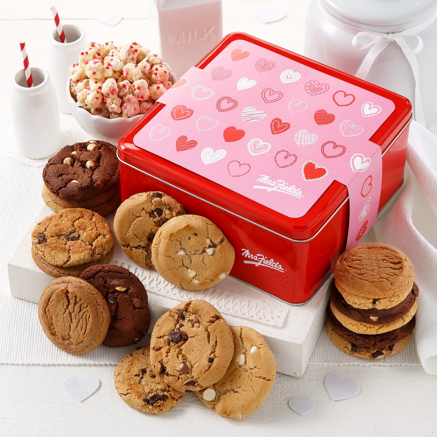 Mrs. Fields signature red cookie tin with a pink Valentine's Day theme sleeve covered in hearts surrounded by an assortment of original cookies and popcorn with heart sprinkles on a white surface.