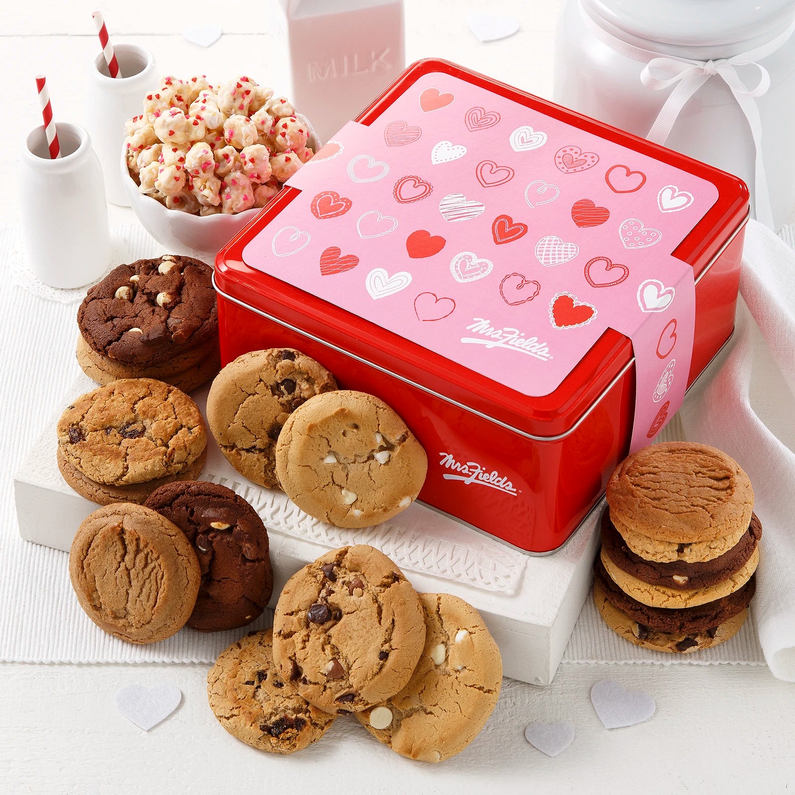 Mrs. Fields signature red cookie tin with a pink Valentine's Day theme sleeve covered in hearts surrounded by an assortment of original cookies and popcorn with heart sprinkles on a white surface.