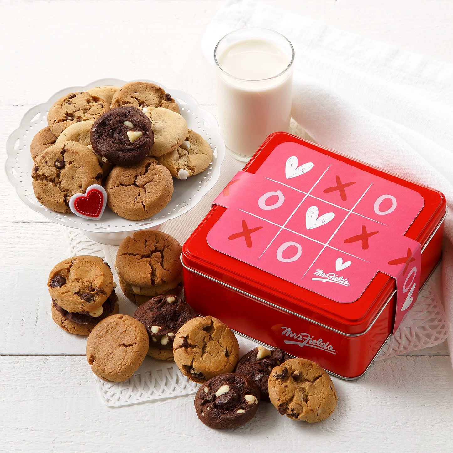 Mrs. Fields signature mini cookie tin with a pink Valentine's Day XOXO tic-tac-toe themed sleeve surrounded an assortment of Nibblers bite-sized cookies on a white surface with a glass of milk.
