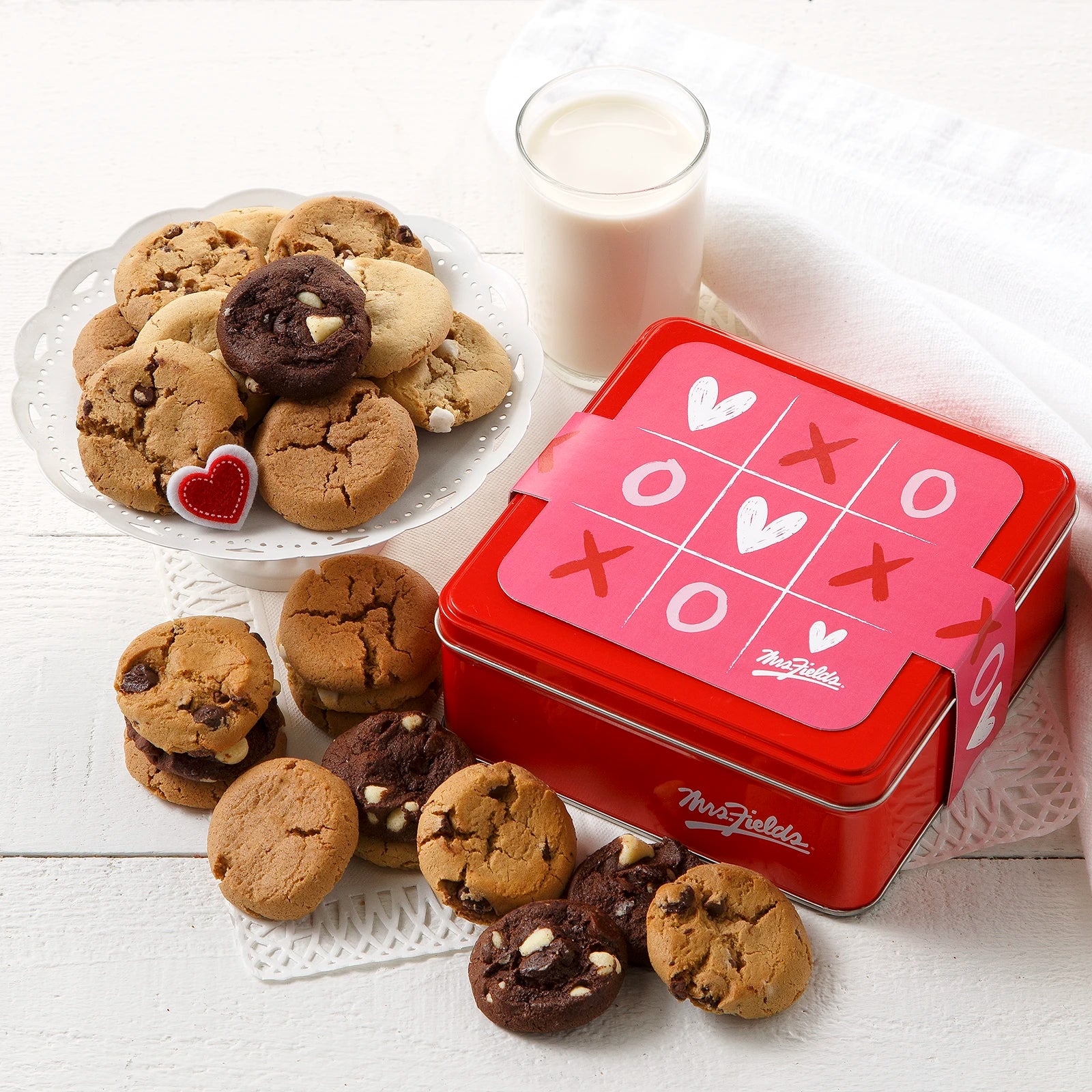 Mrs. Fields signature mini cookie tin with a pink Valentine's Day XOXO tic-tac-toe themed sleeve surrounded an assortment of Nibblers bite-sized cookies on a white surface with a glass of milk.