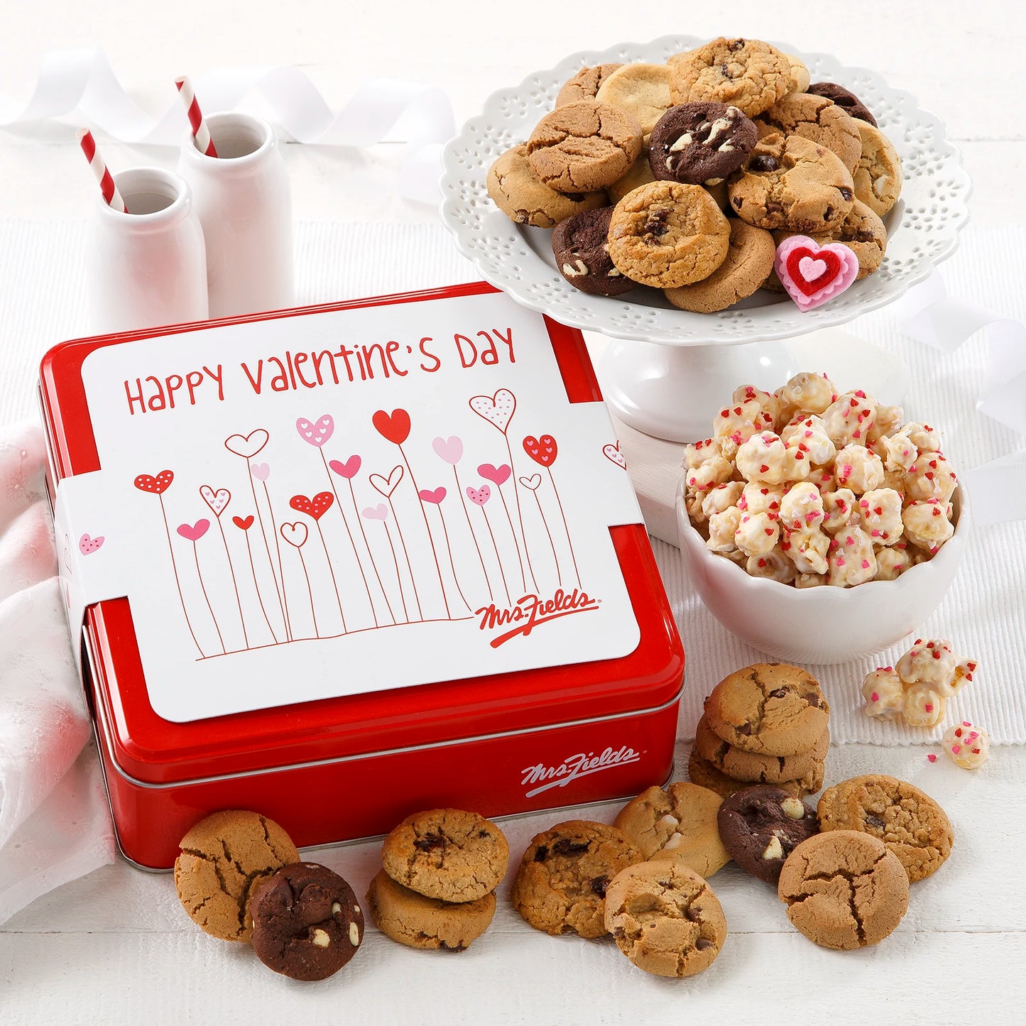 Mrs. Fields signature red cookie tin with a white sleeve that says "Happy Valentine's Day" with hearts on it surrounded by an assortment of Nibblers bite-sized cookies and popcorn with heart sprinkles on a white surface.