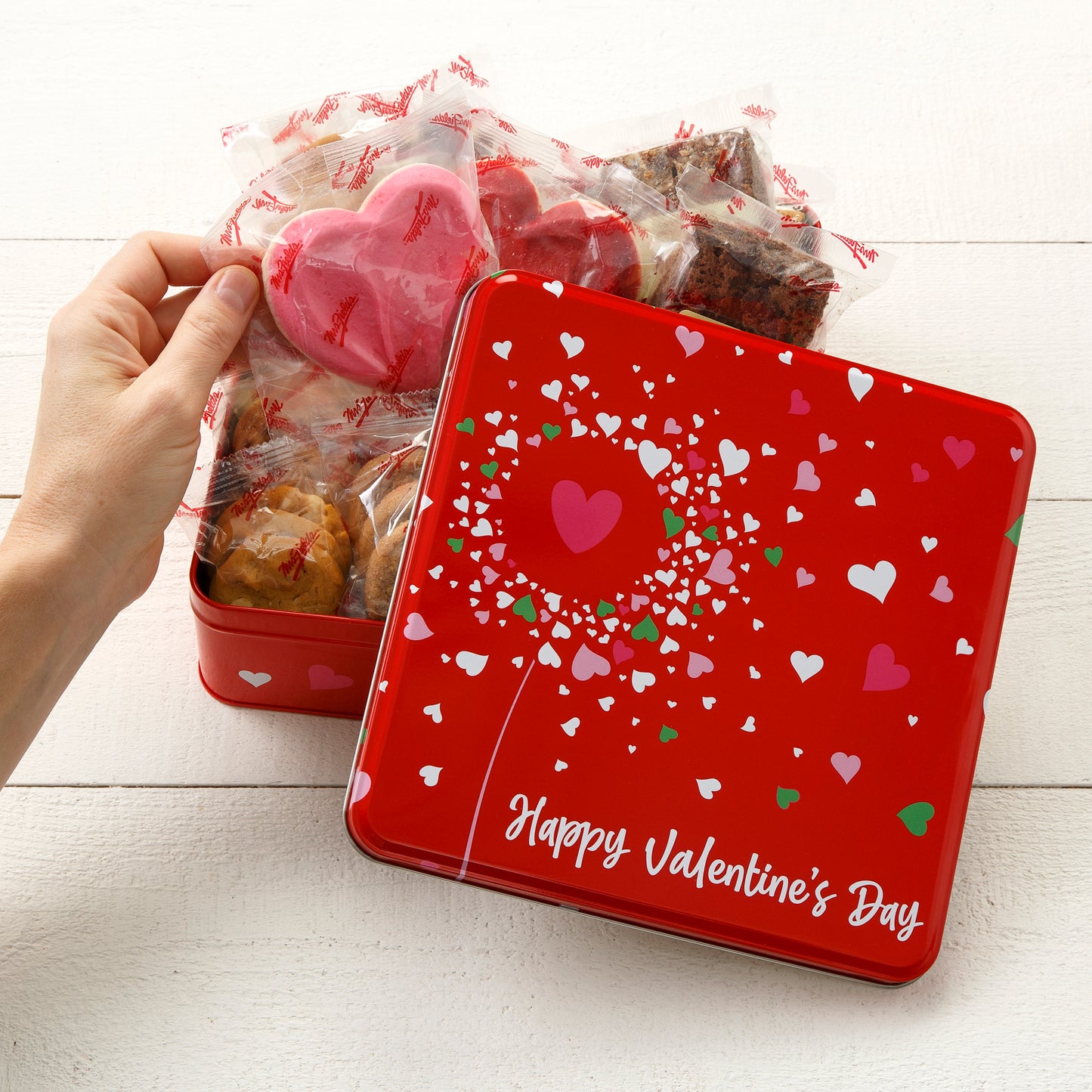 A red with hearts Valentine's Day tin filled with an assortment of packaged nibblers, brownie bites, and frosted heart cookies