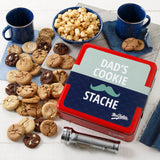 Mrs. Fields signature red cookie tin with a Father's Day themed sleeve that says "Dad's Cookie Stache" with a mustache on it surrounded by an assortment of Nibblers bite-sized cookies and toffee nut popcorn on a white surface with two dark blue mugs and a flashlight.