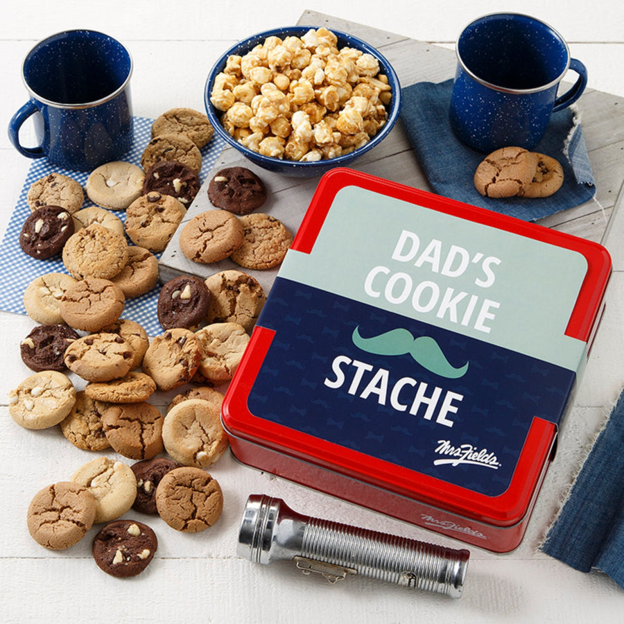 Mrs. Fields signature red cookie tin with a Father's Day themed sleeve that says "Dad's Cookie Stache" with a mustache on it surrounded by an assortment of Nibblers bite-sized cookies and toffee nut popcorn on a white surface with two dark blue mugs and a flashlight.