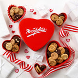 Mrs. Fields signature heart shaped tin surrounded by Nibblers bite-sized cookies in heart shaped dishes on a white surface with felt heart decorations and a red and white ribbon.