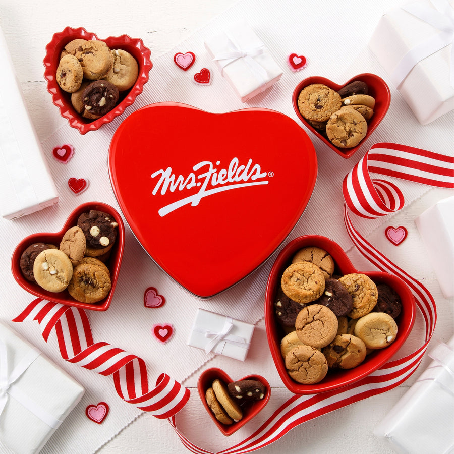 Mrs. Fields signature heart shaped tin surrounded by Nibblers bite-sized cookies in heart shaped dishes on a white surface with felt heart decorations and a red and white ribbon.