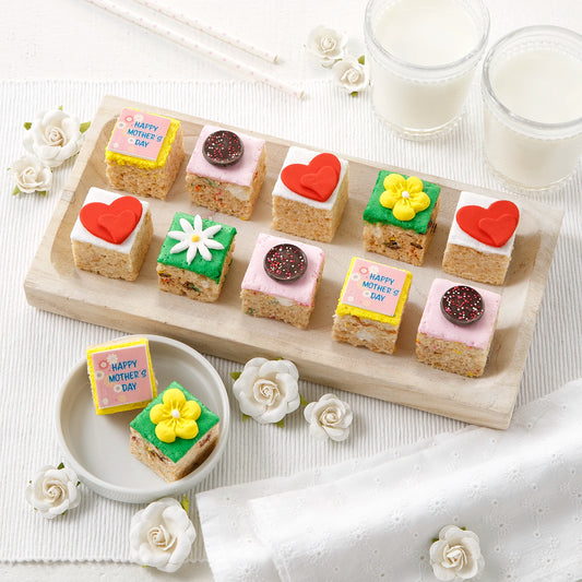 An assortment of twelve mini rice krispie treats all decorated with a springtime and Mother's Day theme with flower and heart decorations, and some that say "Happy Mother's Day" right on them! All on a white surface with white flowers and two glasses of milk.