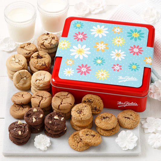 Mrs. Fields signature red tin with a floral spring themed sleeve on it surrounded by an assortment of Nibblers bite-sized cookies on a white surface with two glasses of milk and white flower decorations.