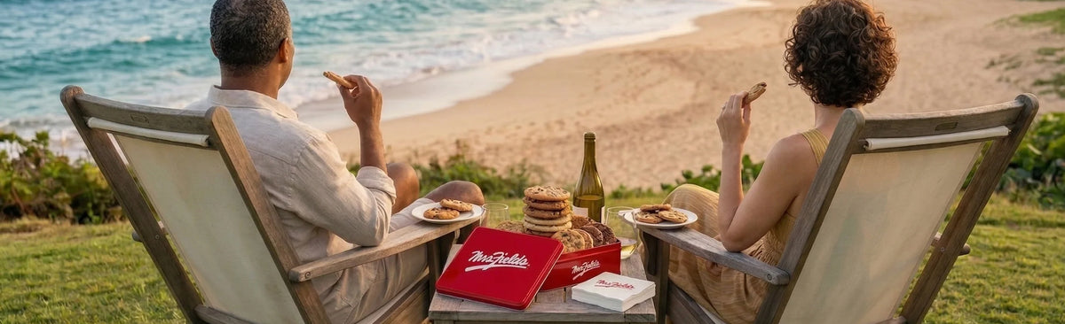 A couple relaxes in wooden beach chairs on a grassy cliff overlooking the ocean, sharing a picnic with a red branded box, plates, and a bottle on a table between them.