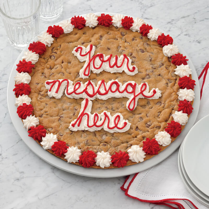 A large chocolate chip cookie cake on a white plate, decorated with red and white whipped frosting around the edge and the words 'Your message here' in red icing in the center, set on a white marble surface with plates and glasses nearby.
