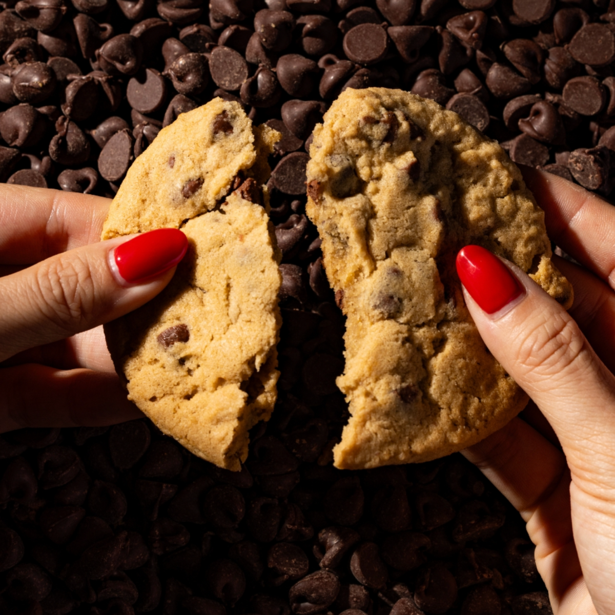 A chocolate chip cookie is broken in half by two hands with bright red painted fingernails, set against a background of dark chocolate chips.