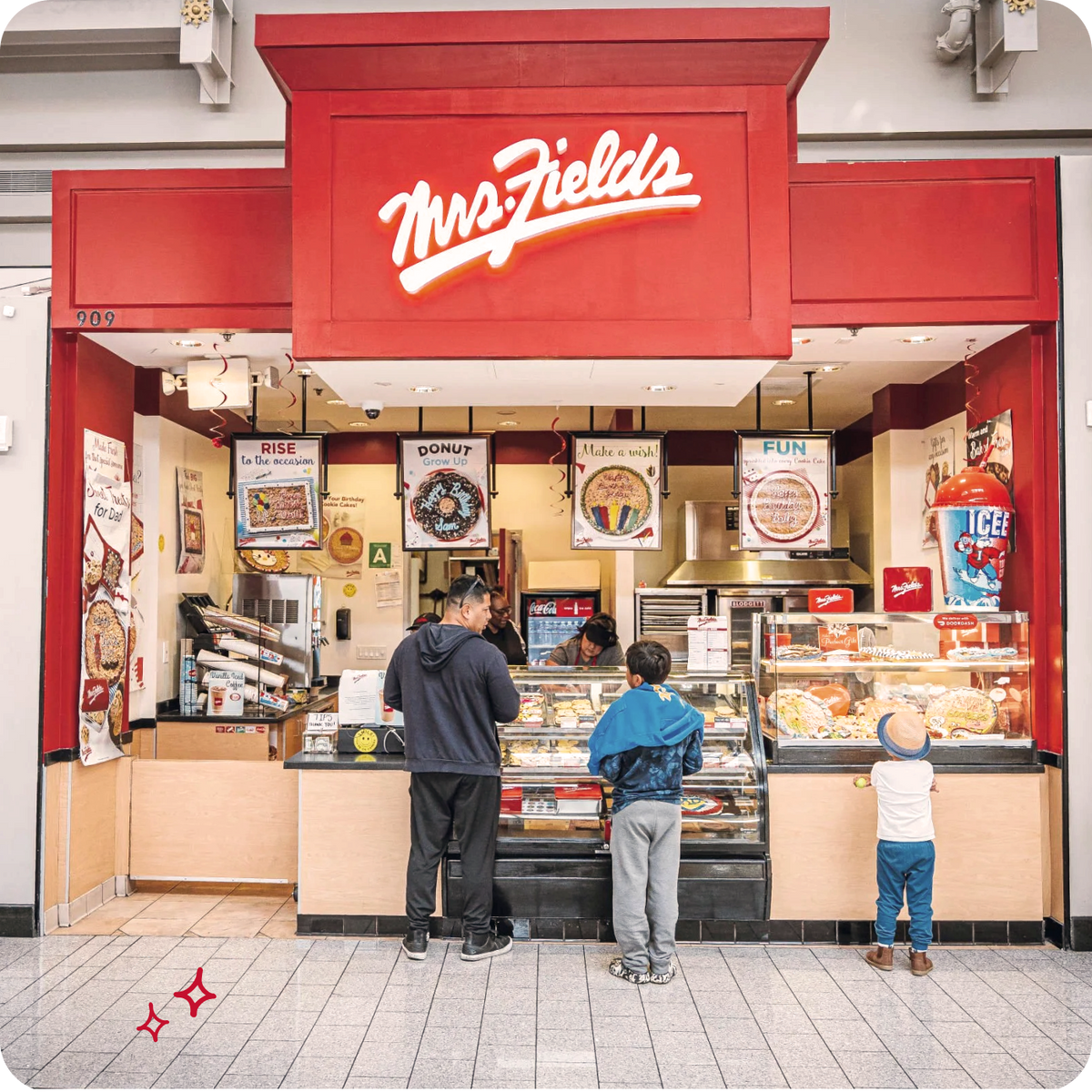 A Mrs. Fields ice cream stand in an indoor shopping center, with three people and a child standing in front of the counter under a red sign.