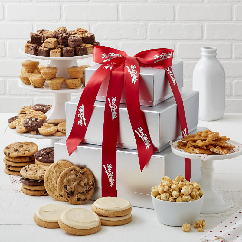 A three tier silver box tower tied with a red Mrs. Fields ribbon and surrounded by an assortment of Nibbers®, original cookies, brownie bites, mini muffins, four frosted cookies, peanut brittle, and popcorn