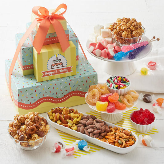 A "Happy Birthday" themed gift tower with reusable boxes surrounded by an assortment of snacks and candy on a white surface.