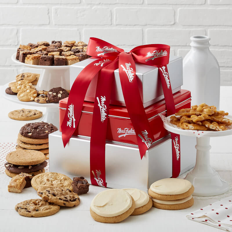 A three tier silver box and red tin tower and tied with a red Mrs. Fields ribbon and surrounded by an assortment of Nibblers®, original cookies, brownie bites, four frosted cookies, and peanut brittle